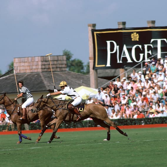 Two polo players on horseback gallop across a green field during a match, with a large "PIAGET" banner and a crowd of spectators in the background
