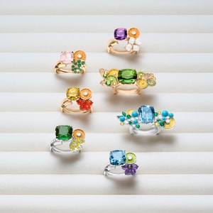 Several Piaget Cocktail rings in rose and white gold, adorned with colorful ornamental stones and stacked designs, presented on a textured white stand.