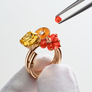 Rose gold cocktail ring with a large yellow stone, a circular orange element, and red motifs, held by a gloved hand, with a red bead being manipulated by tweezers.