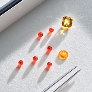 Composition of red beads, a faceted yellow citrine, a carved yellow stone, and tweezers, presented on a textured white background.