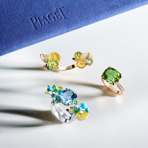 Three colorful Piaget Cocktail rings, including one in white gold with a blue aquamarine and floral motifs, and one in rose gold with a tourmaline stone, placed on a white surface. A blue case marked 'PIAGET' is visible in the background.